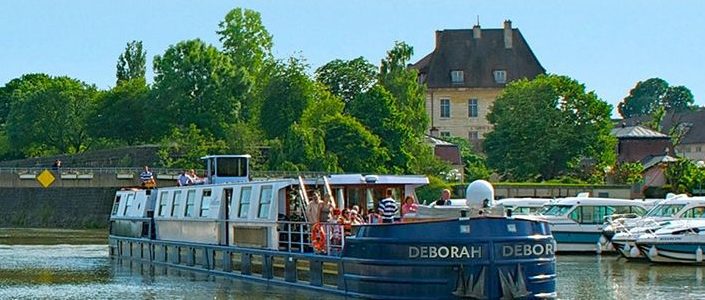 Hotel barge Deborah on a river in France with a historic building and some greenery in the background